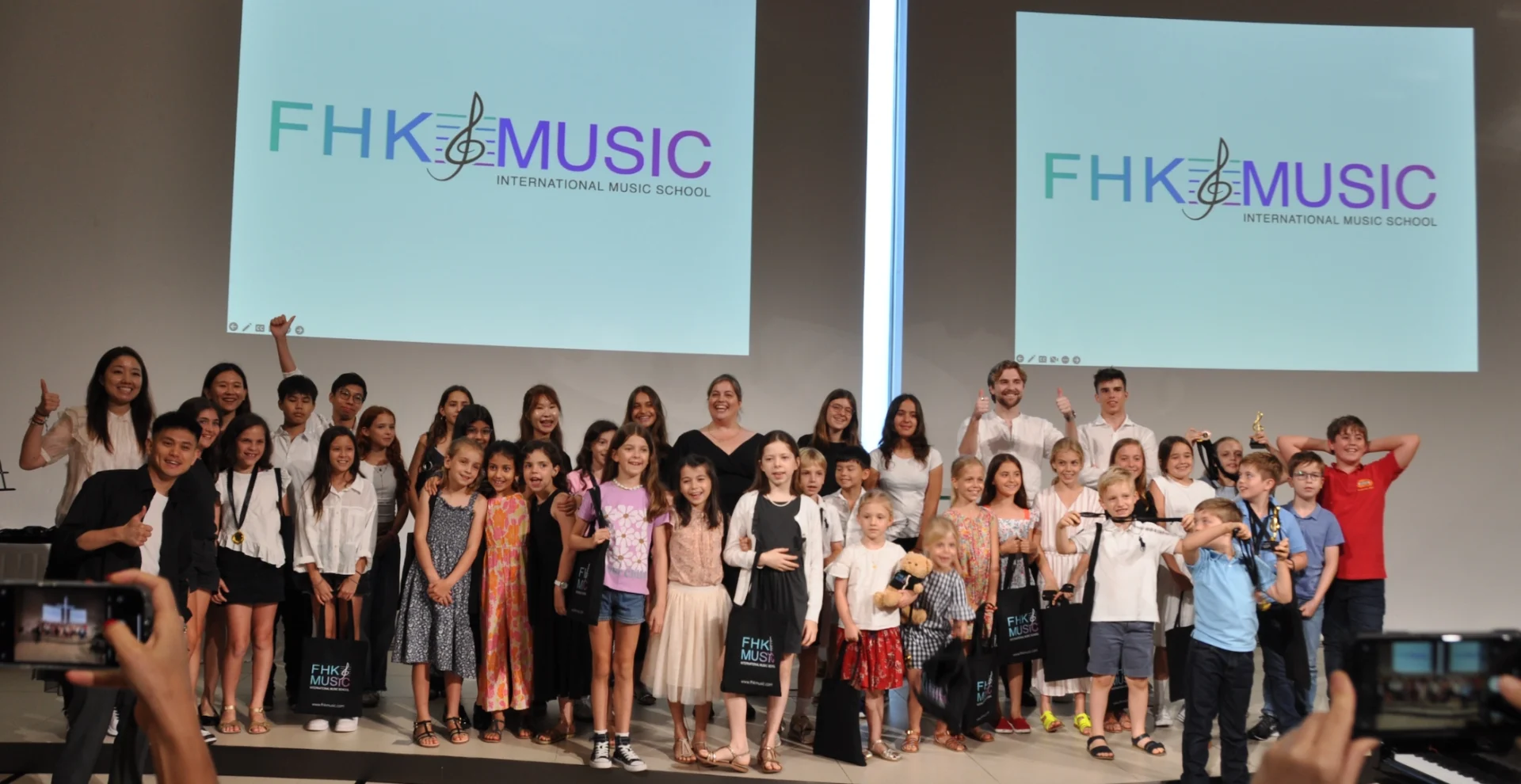 Group of FHK Music students smiling on stage with the FHK Music International Music School logo projected behind them
